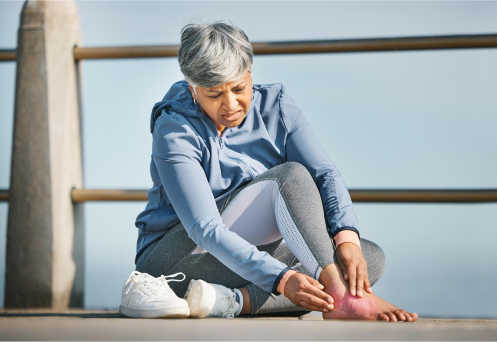 Older woman sitting outdoors, holding her foot in pain, possibly experiencing discomfort that may require a podiatrist's attention.