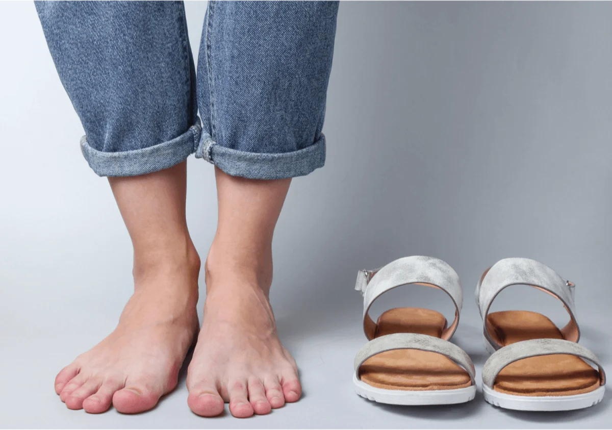 A pair of bare feet standing next to silver sandals on a clean background.